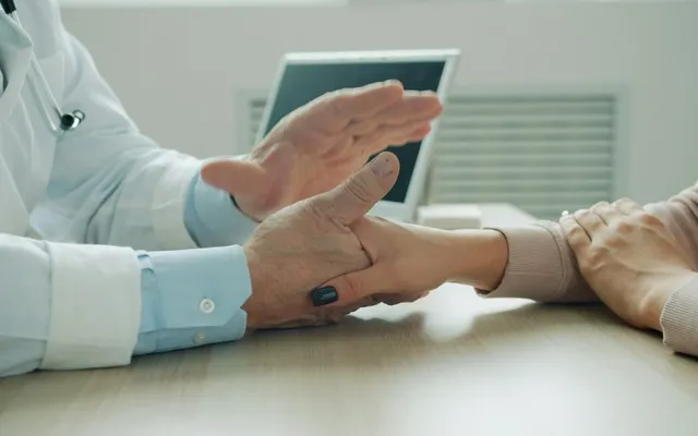 Physician in a white coat holding a patient's hand at a light desk, with a dark-screen laptop in the background