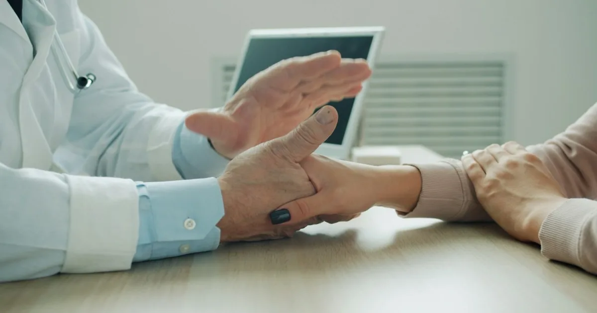 Physician in a white coat holding a patient's hand at a light desk, with a dark-screen laptop in the background